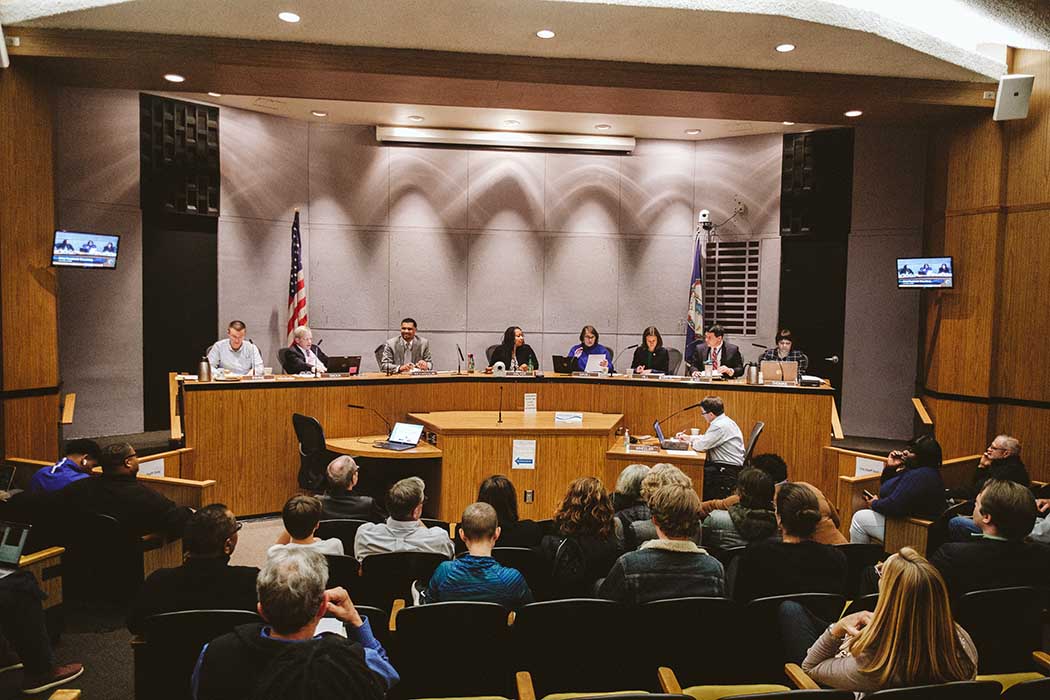 Far away photo of Charlottesville City Councilors sitting on the dais during a pre-COVID meeting inside City Hall. Around a dozen people sit in the audience, facing the councilors