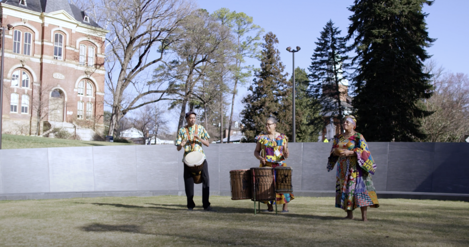 Two Black woman and one Black man play traditional drums on a green lawn in front the granite walls of the Memorial to Enslaved Laborers.